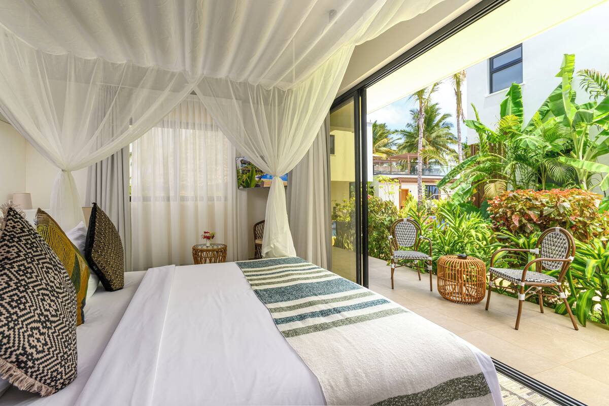 A bedroom with a large bed, white curtains, and a view of a tropical garden through a glass door.