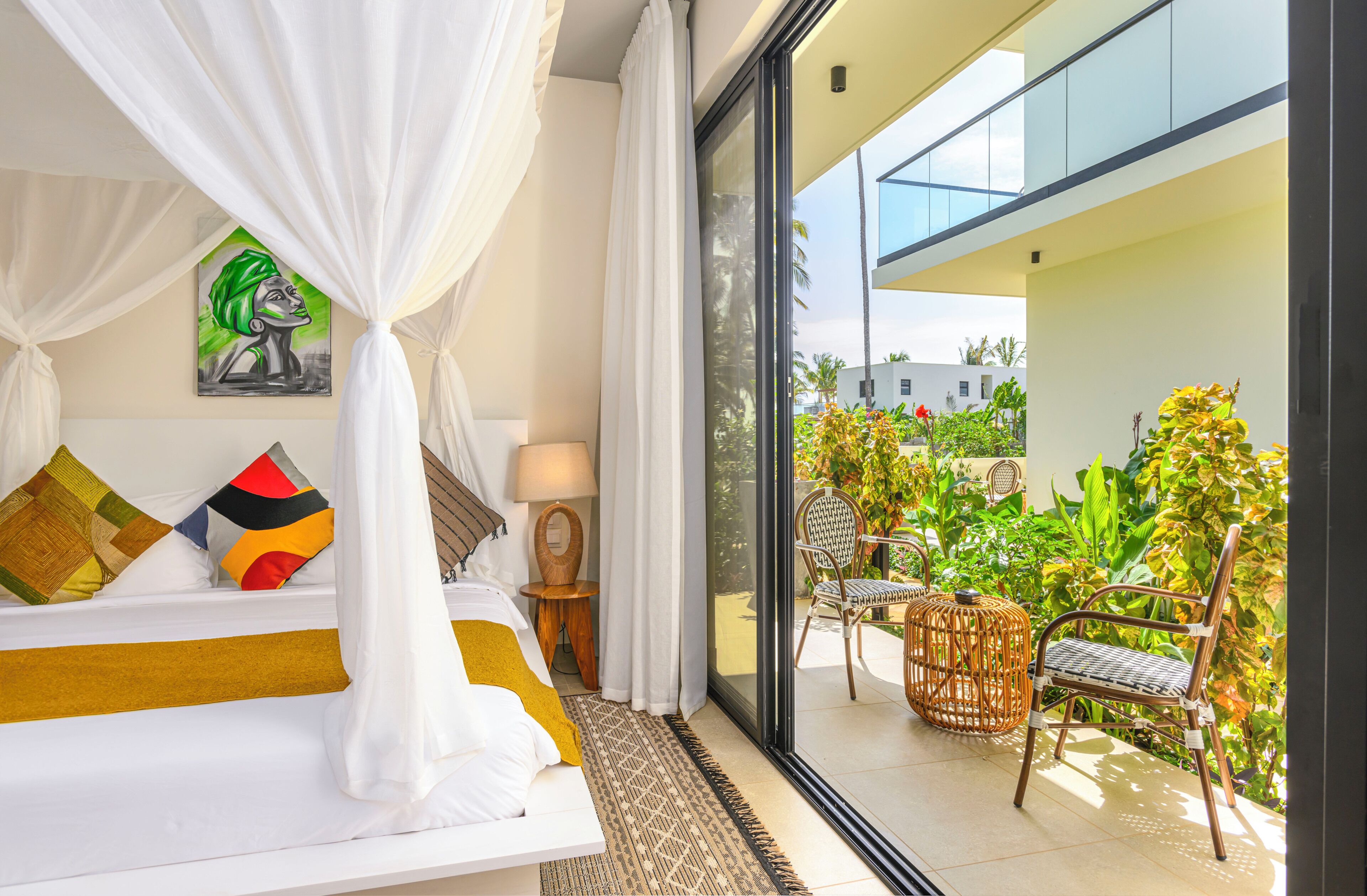 A bedroom with a bed, a nightstand, a lamp, a painting, and a view of the balcony with chairs and plants.