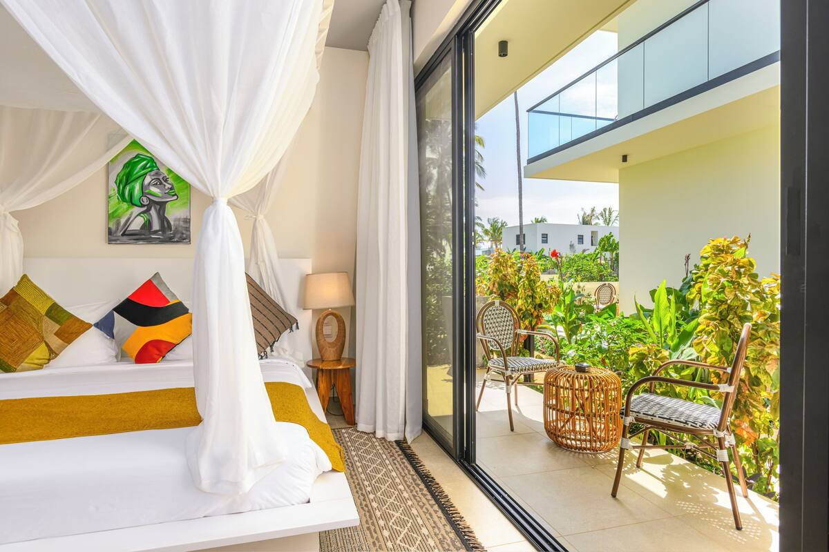 A bedroom with a bed, a nightstand, a lamp, a painting, and a view of the balcony with chairs and plants.