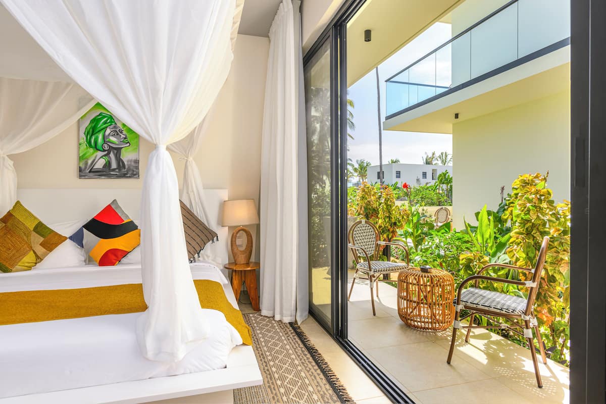 A bedroom with a bed, a nightstand, a lamp, a painting, and a view of the balcony with chairs and plants.