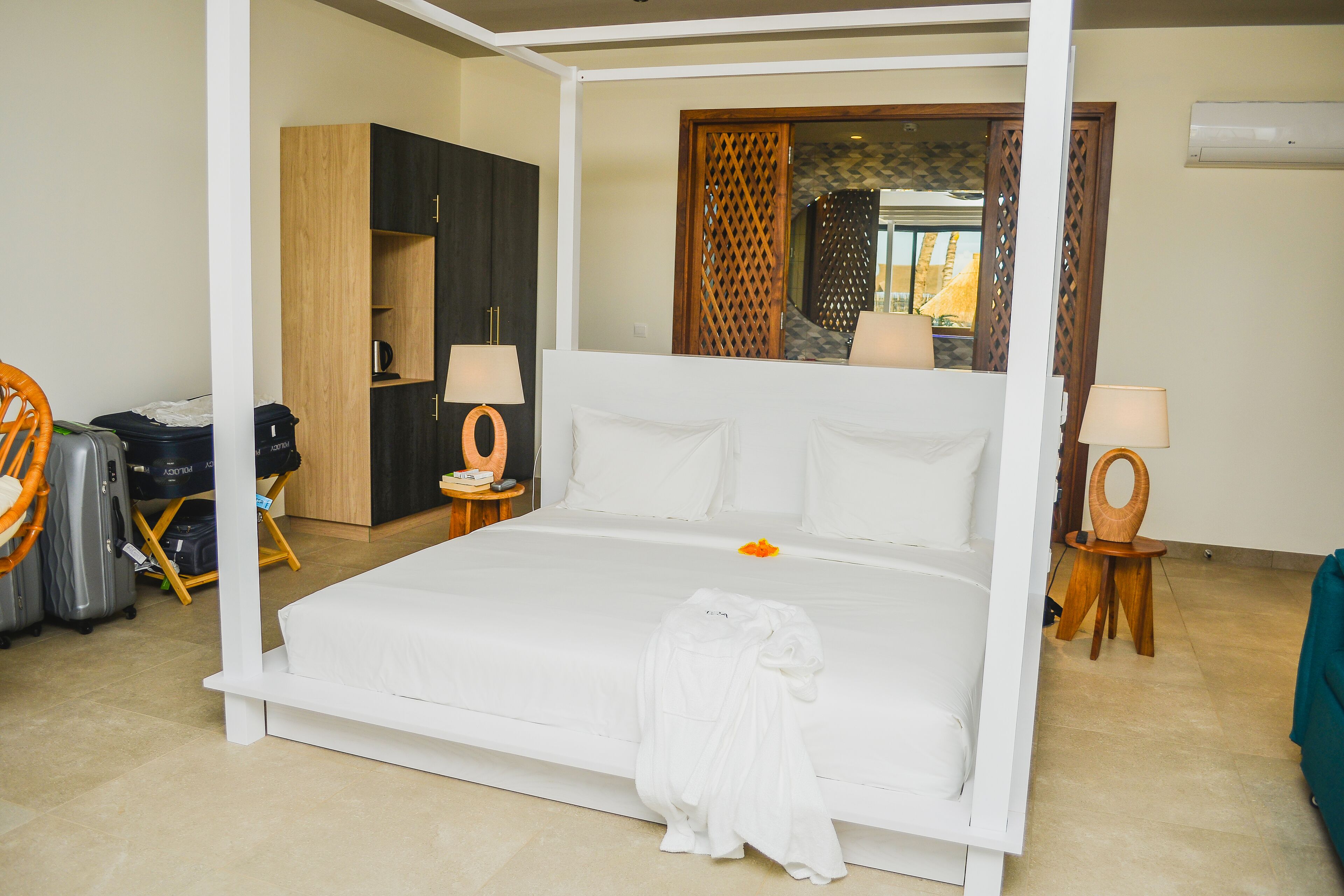 A four-poster bed with white linens, a wooden wardrobe, a small round table with a lamp, and a wicker chair.