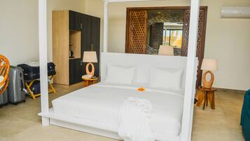 A four-poster bed with white linens, a wooden wardrobe, a small round table with a lamp, and a wicker chair.
