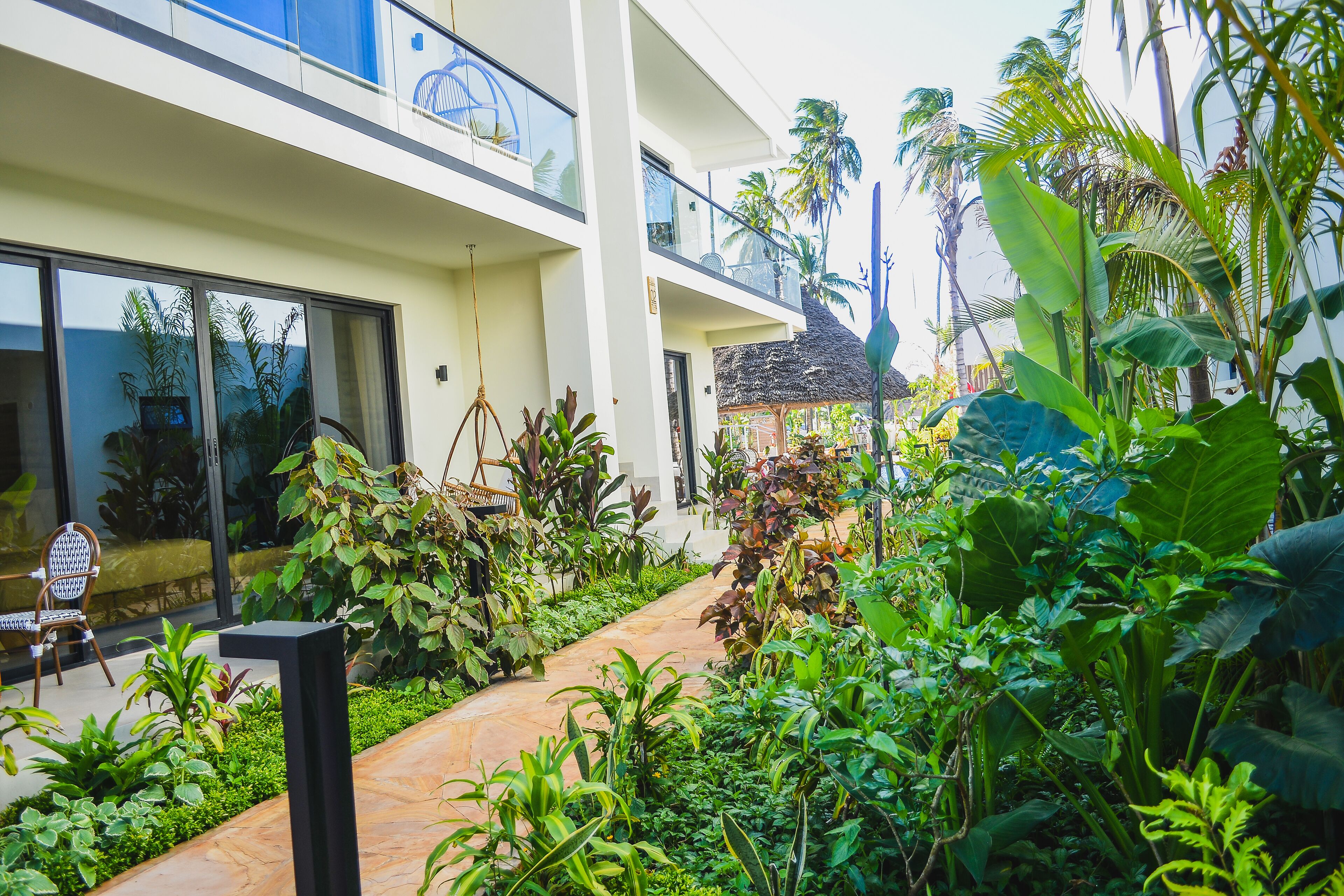 Um edifício moderno com varanda de vidro, cercado por plantas tropicais exuberantes e um caminho pavimentado.