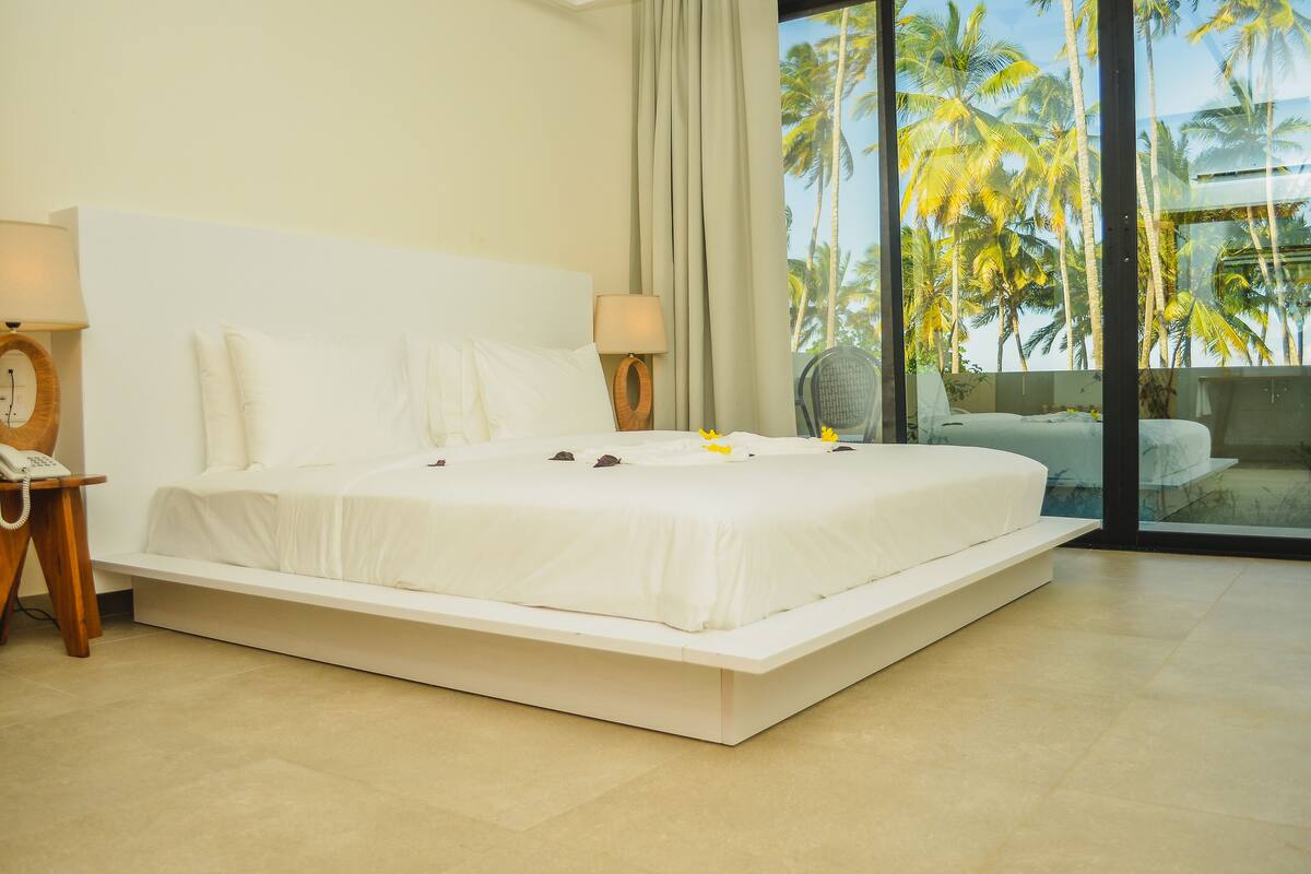 A modern hotel room with a large bed, a bedside telephone, and a view of palm trees through large windows.