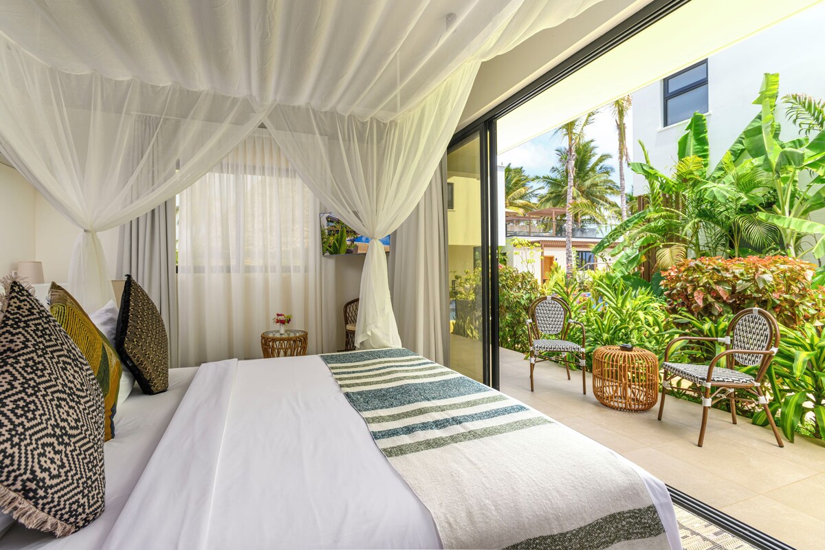 Un dormitorio con una cama grande, cortinas blancas y vista a un jardín tropical a través de un ventanal.