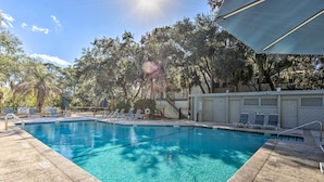 En klar swimmingpool med fliseterrasse, omgivet af træer og et overdækket område med liggestole.