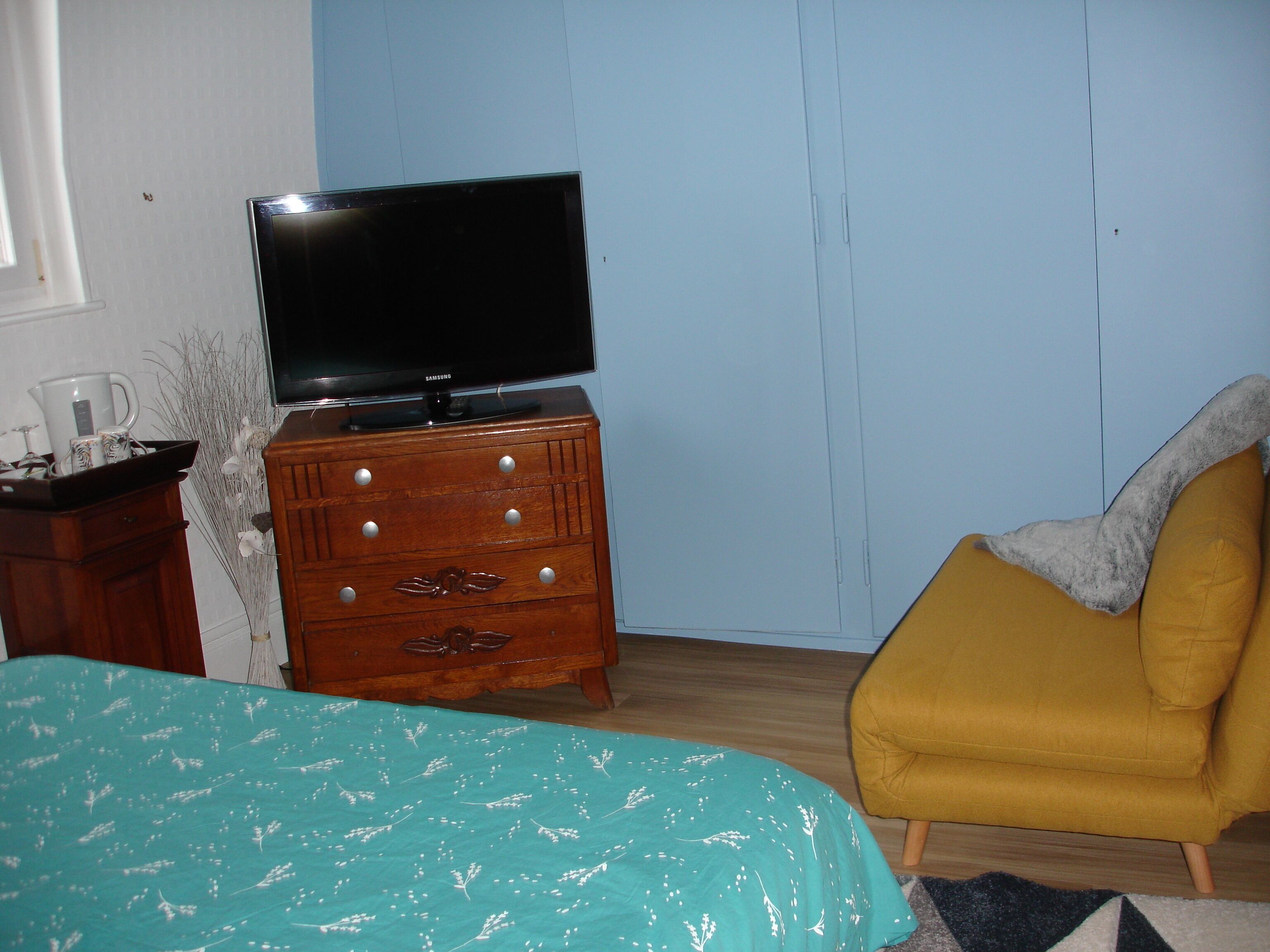 Un dormitorio con una cama, un aparador de madera con televisión de pantalla plana, un sillón amarillo y una ventana con persianas.