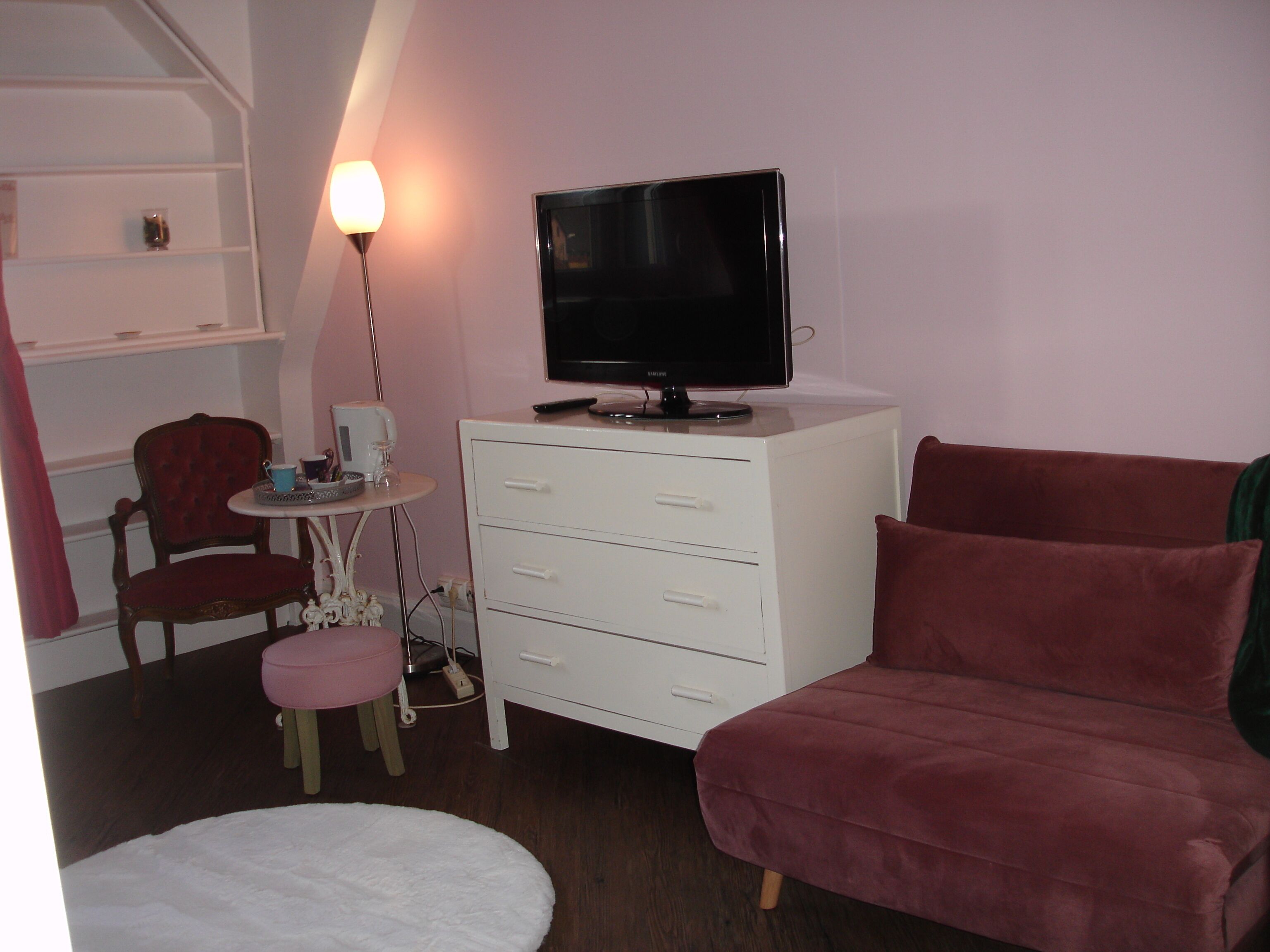 Ein kleines Zimmer mit Fernseher auf einer weißen Kommode, einem roten Sessel, einem kleinen runden Tisch mit Lampe und einem pinkfarbenen Hocker.