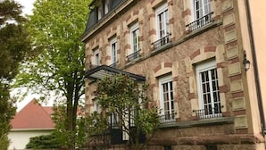 A two-story stone building with multiple windows and balconies, surrounded by a well-maintained garden with potted plants and a wooden fence. - Les Jardins d'Aïka (Raon-l'Etape)