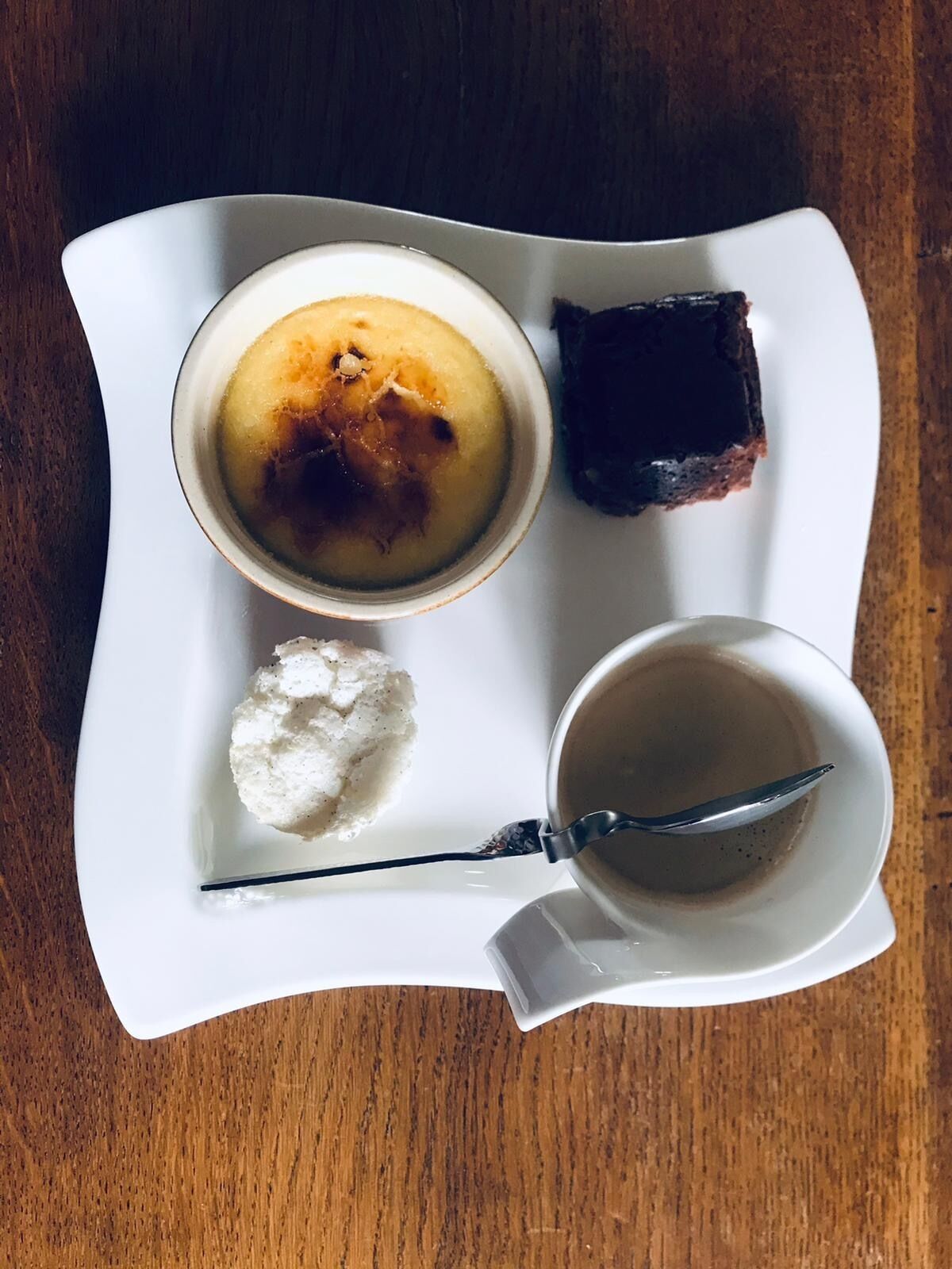 A white plate with a dessert consisting of a caramelized top, a piece of dark chocolate, a scoop of ice cream, and a cup of coffee.
