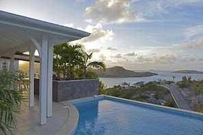 A swimming pool with a view of the sea and mountains, a covered outdoor area with plants, and a stone wall. - Villa LAGON JAUNE – Twin Villa Estate with 7 Bedrooms & Ocean Views in Petit Cul de Sac, St. Barths (Gustavia)