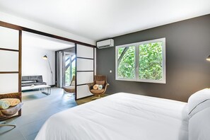 Quarto de hotel moderno com uma cama grande, área de estar com sofá e poltrona, vista para o verde pela janela e um aparelho de ar condicionado na parede.