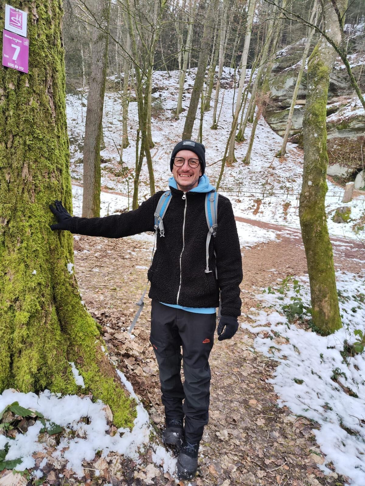 A person standing next to a moss-covered tree in a forested area with snow on the ground.
