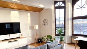 Een moderne woonkamer met een flatscreen televisie, een grijze bank, een houten vloer, grote ramen en uitzicht op de stad.