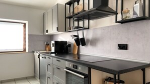 Een moderne keuken met grijze kastjes, een zwart aanrechtblad en een ingebouwde oven. Er is een raam dat natuurlijk licht biedt, en een plank met keukengerei.