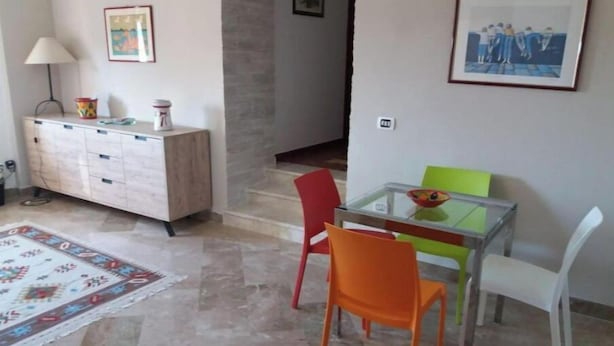 A dining area with a glass table, colorful chairs, a white chair, a patterned rug, a wooden cabinet, and framed pictures on the walls. - Miscale House (Cattolica Eraclea)