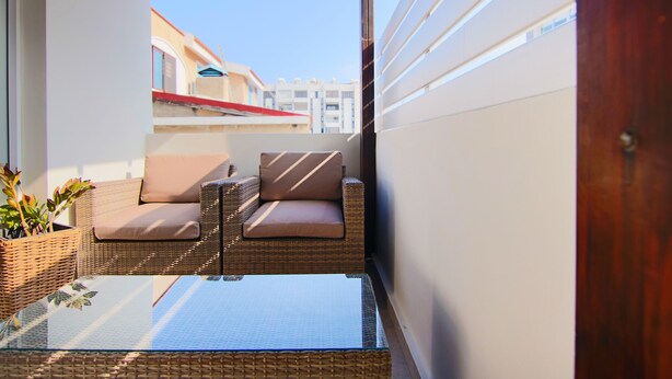 A balcony with wicker furniture and a glass table, overlooking a building with a red roof. - City View Luxury Flat Harbour 04 (Larnaca)