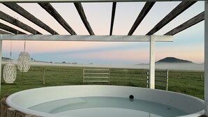 Un jacuzzi grande al aire libre bajo una pérgola de madera, con vista a un paisaje brumoso y colinas distantes.