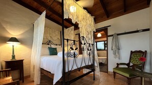 Una cama con dosel y cortinaje, una silla de madera, una mesita de noche y vistas a un baño.
