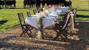 Un área de comedor al aire libre con mesas preparadas para los huéspedes, rodeada por un campo de césped y una manada de vacas al fondo.