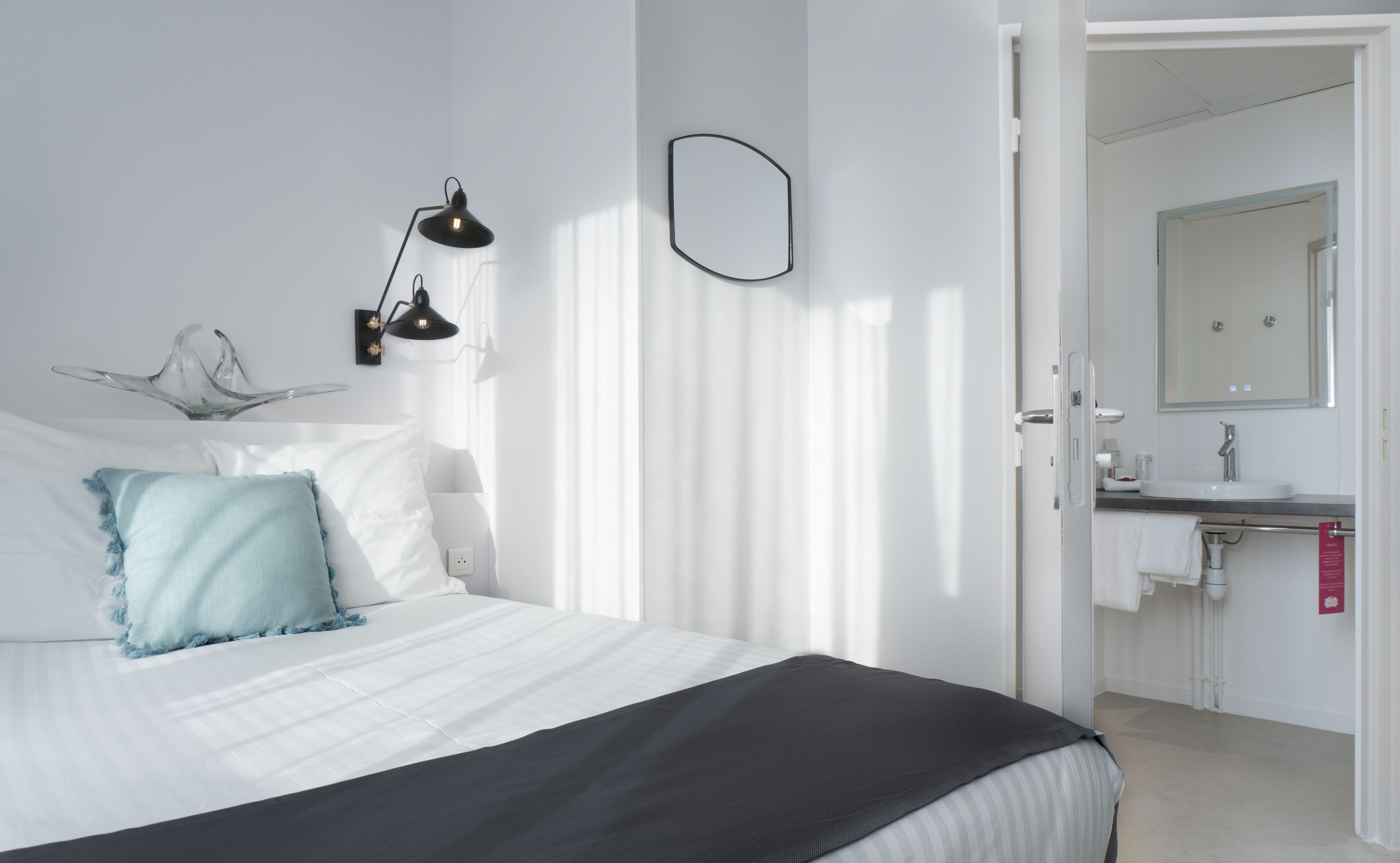 A bedroom with a bed, two wall-mounted lamps, a decorative glass piece, and a bathroom with a mirror and sink.