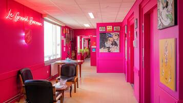 A brightly colored hallway with a neon sign reading "Le Grand Barossan" and a shield emblem. There are chairs and tables along the walls, and artwork is displayed on the walls.