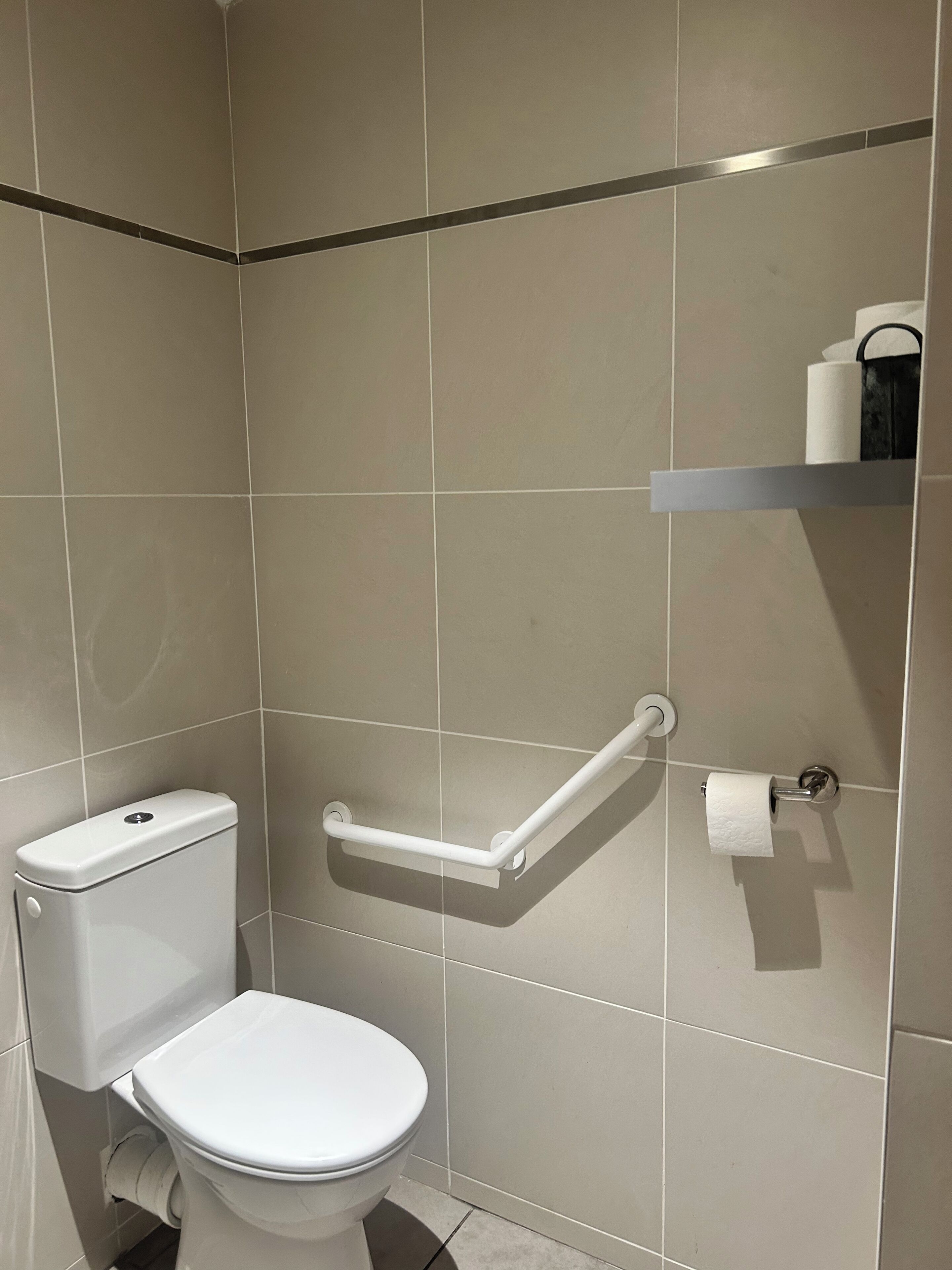 Un baño con inodoro, barra de apoyo y estante con artículos de aseo.
