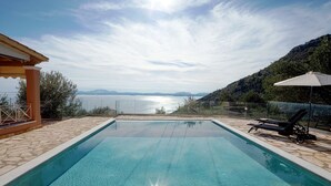 Una piscina rectangular con agua azul cristalina, rodeada de baldosas de piedra. Hay sillones de descanso y una sombrilla a la derecha. La piscina tiene como fondo un cielo despejado y una vista distante de una masa de agua y colinas.