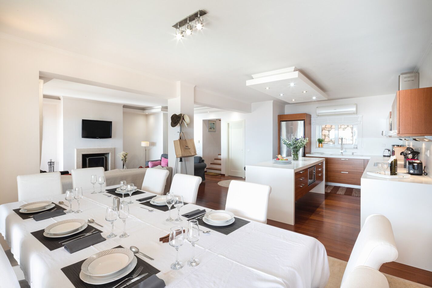 Un comedor moderno con una mesa grande dispuesta para una comida, una zona de cocina con armarios de madera, y un espacio de sala de estar con chimenea.