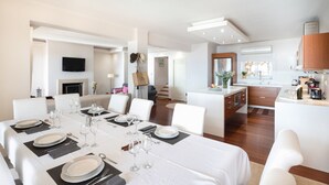 Un comedor moderno con una mesa grande dispuesta para una comida, una zona de cocina con armarios de madera, y un espacio de sala de estar con chimenea.