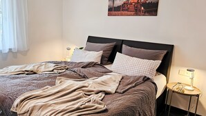Ein ordentlich bezogenes Bett mit einem karierten Kopfkissen, ein Nachttisch mit einer Lampe und ein Gemälde einer Ernteszene an der Wand.