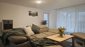 Ein modernes Wohnzimmer mit einem beigefarbenen Sofa, einem Holztisch und einem kleinen Beistelltisch. Auf dem Tisch steht eine Pflanze und eine Vase mit Blumen.
