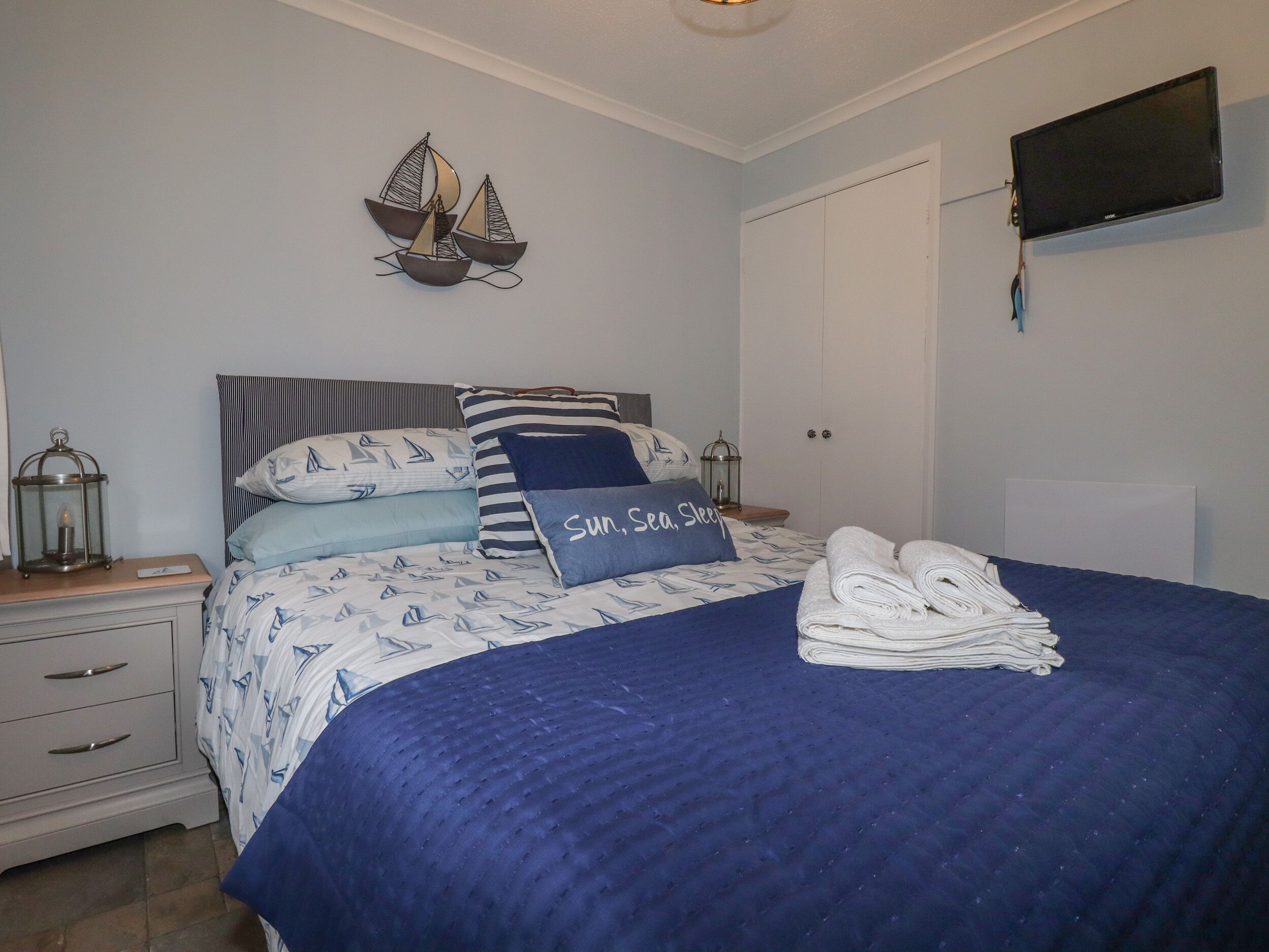 A neatly made bed with a blue and white nautical-themed bedspread, a pillow with "Sun, Sea, Sleep" text, and a sailboat wall decoration.