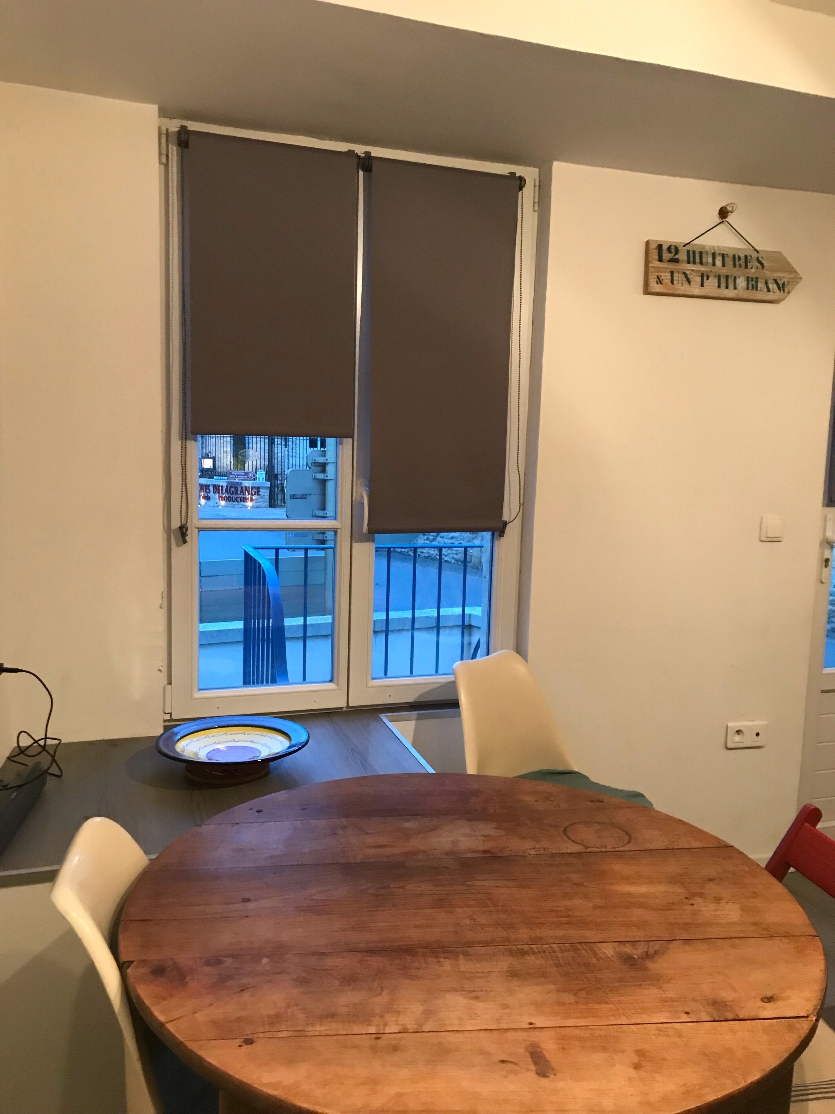 Een kamer met een houten tafel, witte stoelen, een raam met jaloezieën en een bord aan de muur.