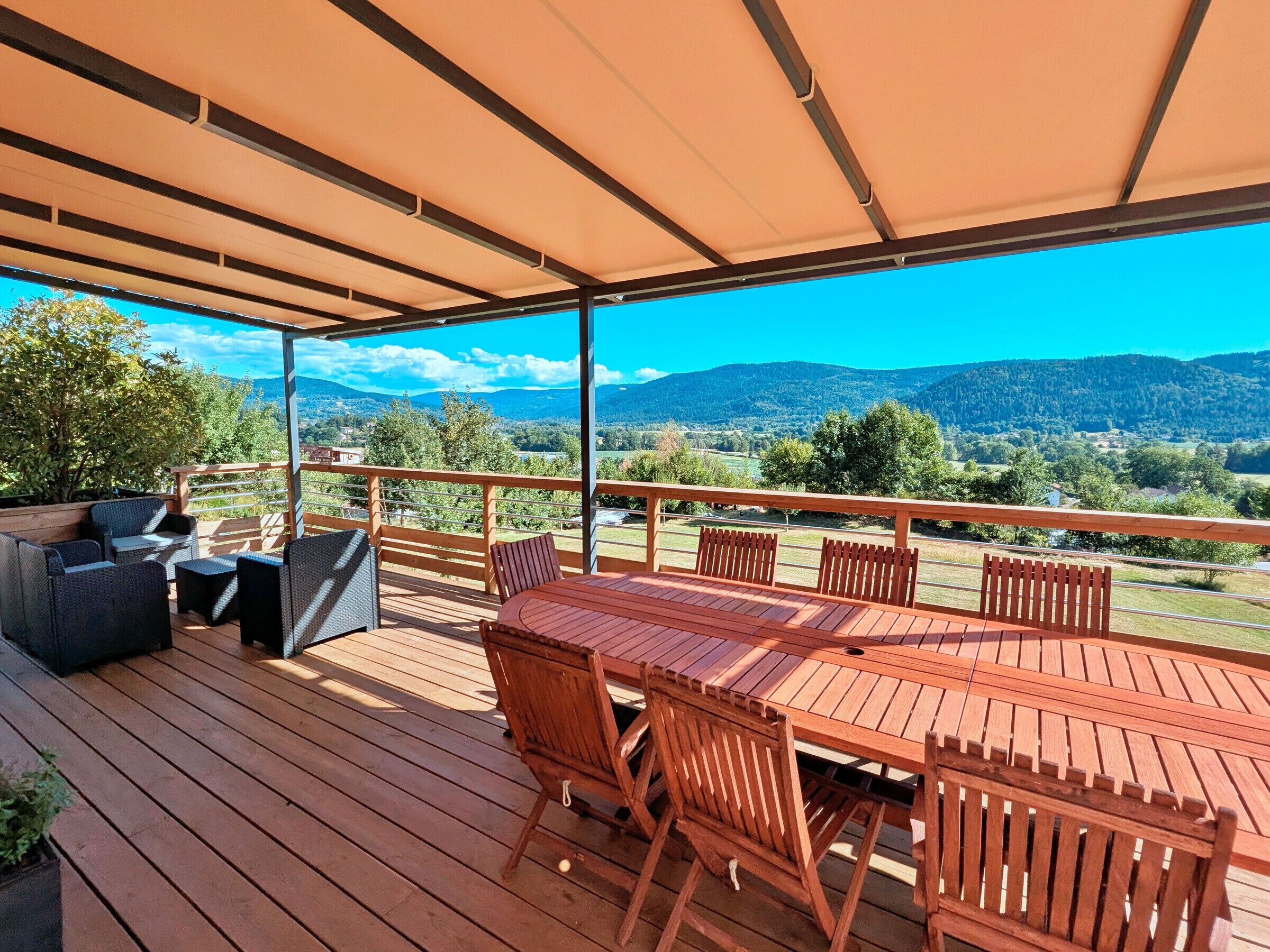 Une terrasse extérieure couverte, dotée d’une table et de chaises en bois, d’un canapé et offrant une vue sur un paysage montagneux.