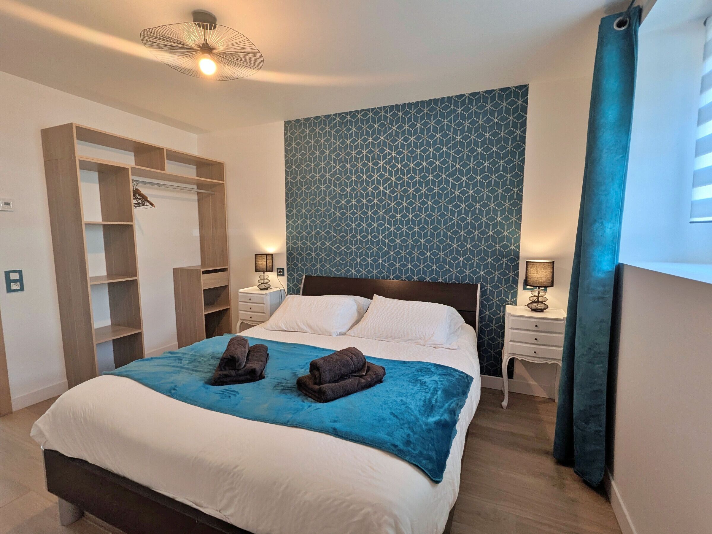 A neatly made bed with a blue blanket, a wooden wardrobe, and a patterned wall.