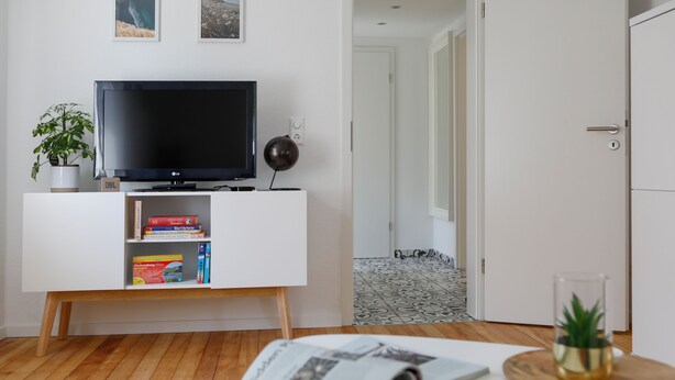 Uma sala de estar moderna com uma TV de tela plana em um armário branco, uma planta pequena e uma revista sobre uma mesa.