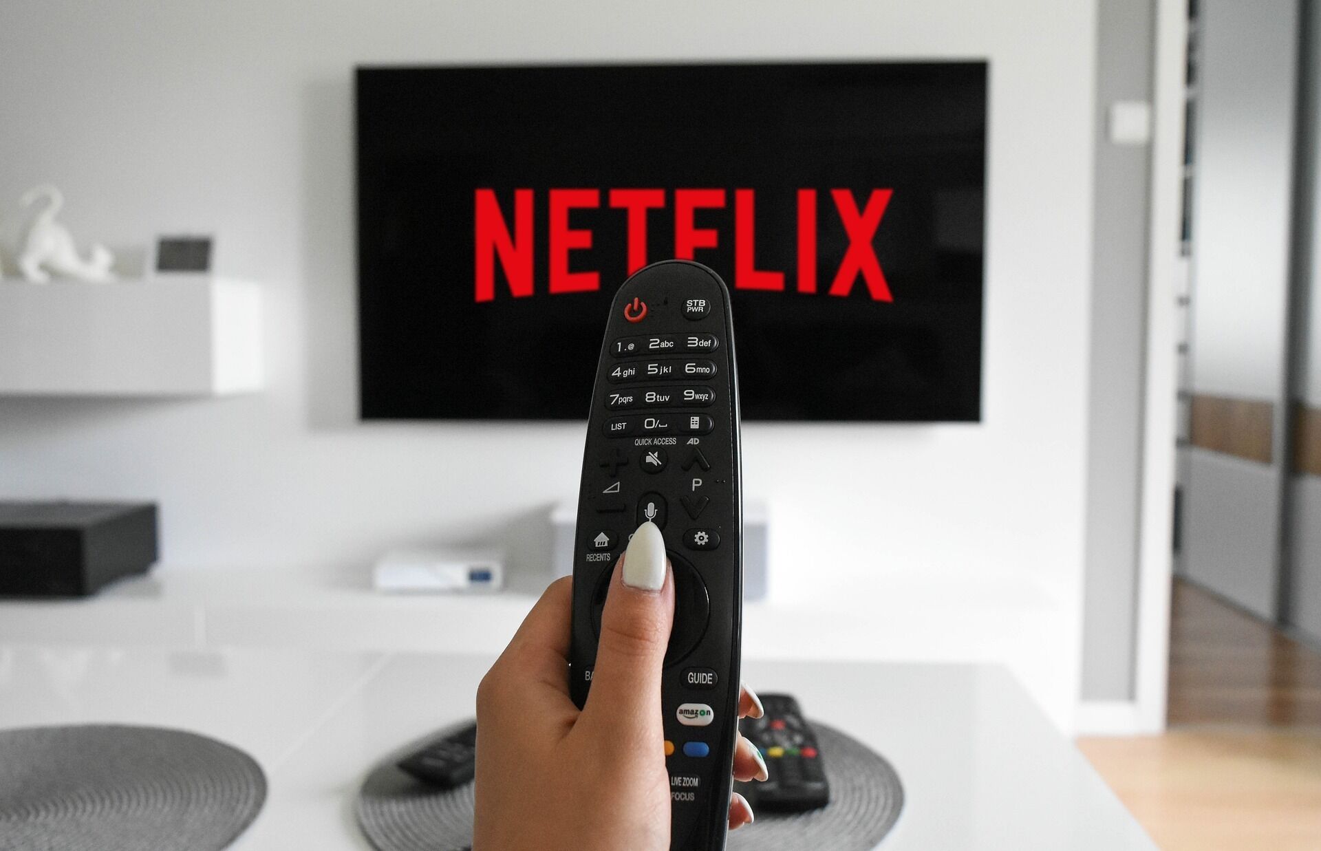 A hand holding a remote control in front of a TV displaying the Netflix logo.
