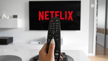 A hand holding a remote control in front of a TV displaying the Netflix logo.