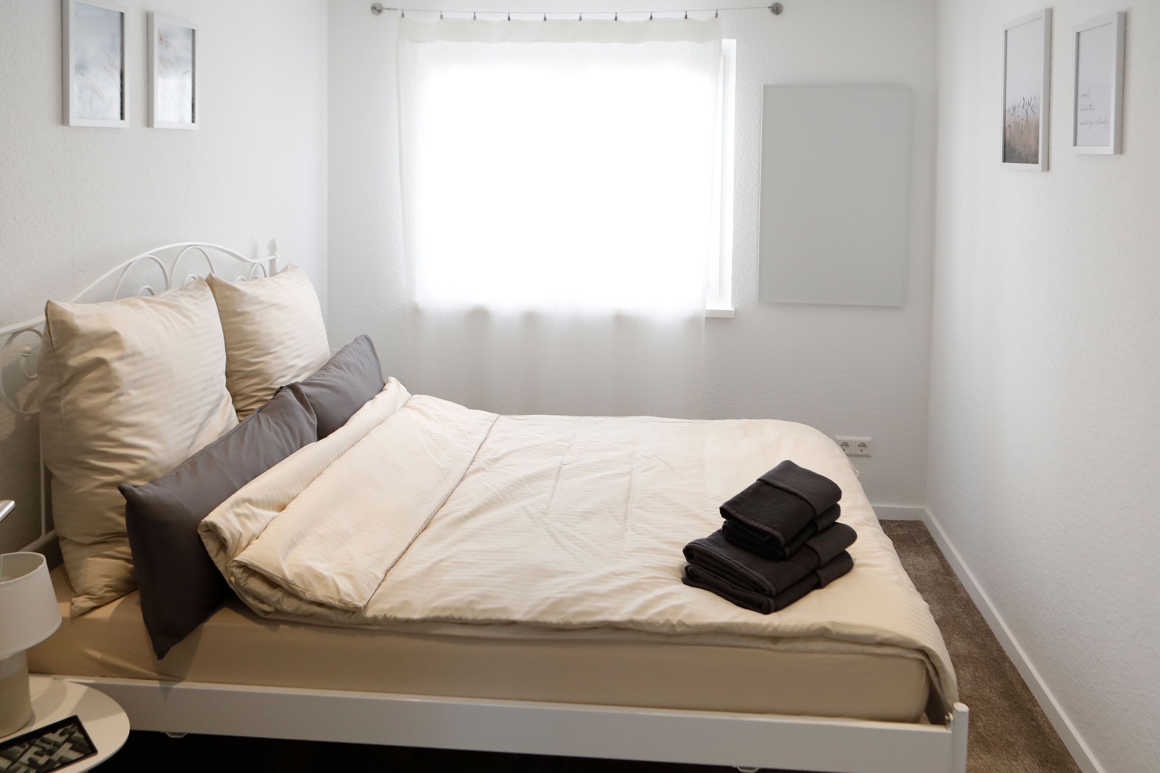 A neatly made bed with a white headboard, a window with sheer curtains, and a small bedside table with a lamp.