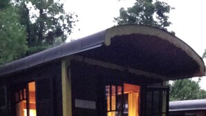 Une petite maison aux couleurs jaune et noir, décorée sur le thème des trains, avec un porche et des marches. À l’intérieur, on trouve une table entourée de chaises où sont assis deux enfants et un adulte.