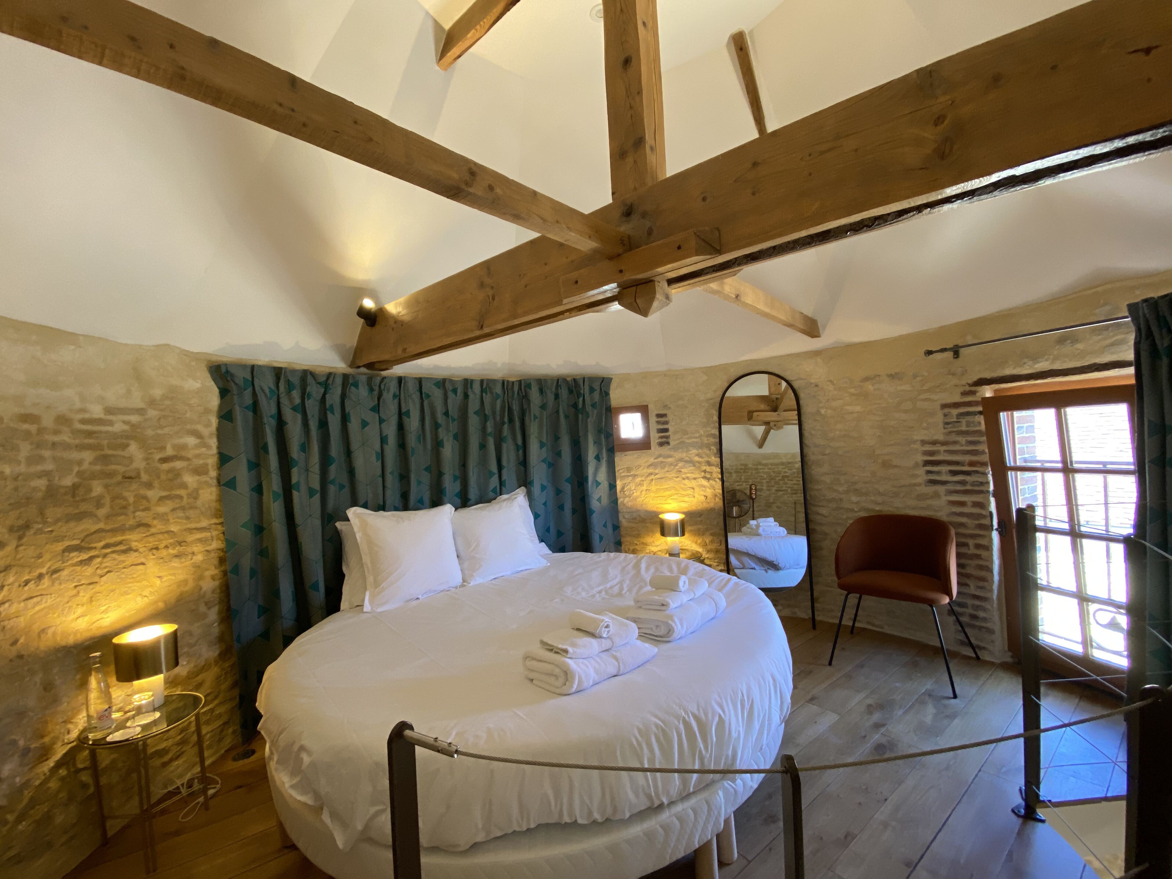 A bedroom with a large bed, wooden beams, stone walls, a mirror, a chair, and a window.
