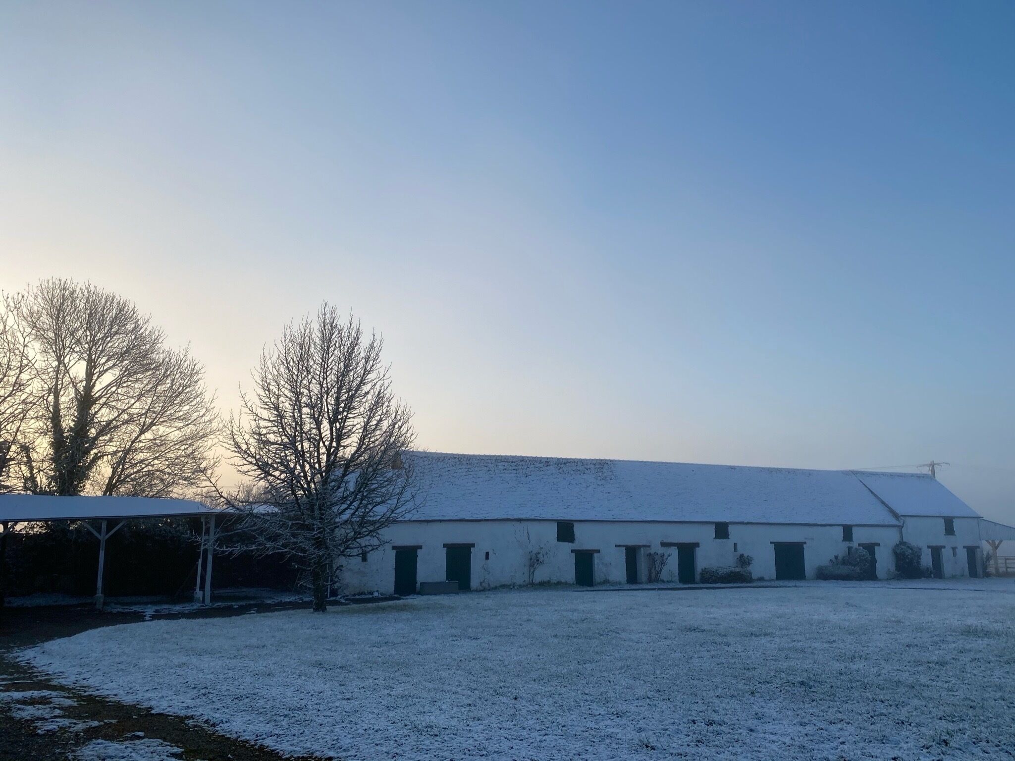 A snow-covered barn with a flat roof and multiple doors, surrounded by a snow-covered field and leafless trees.