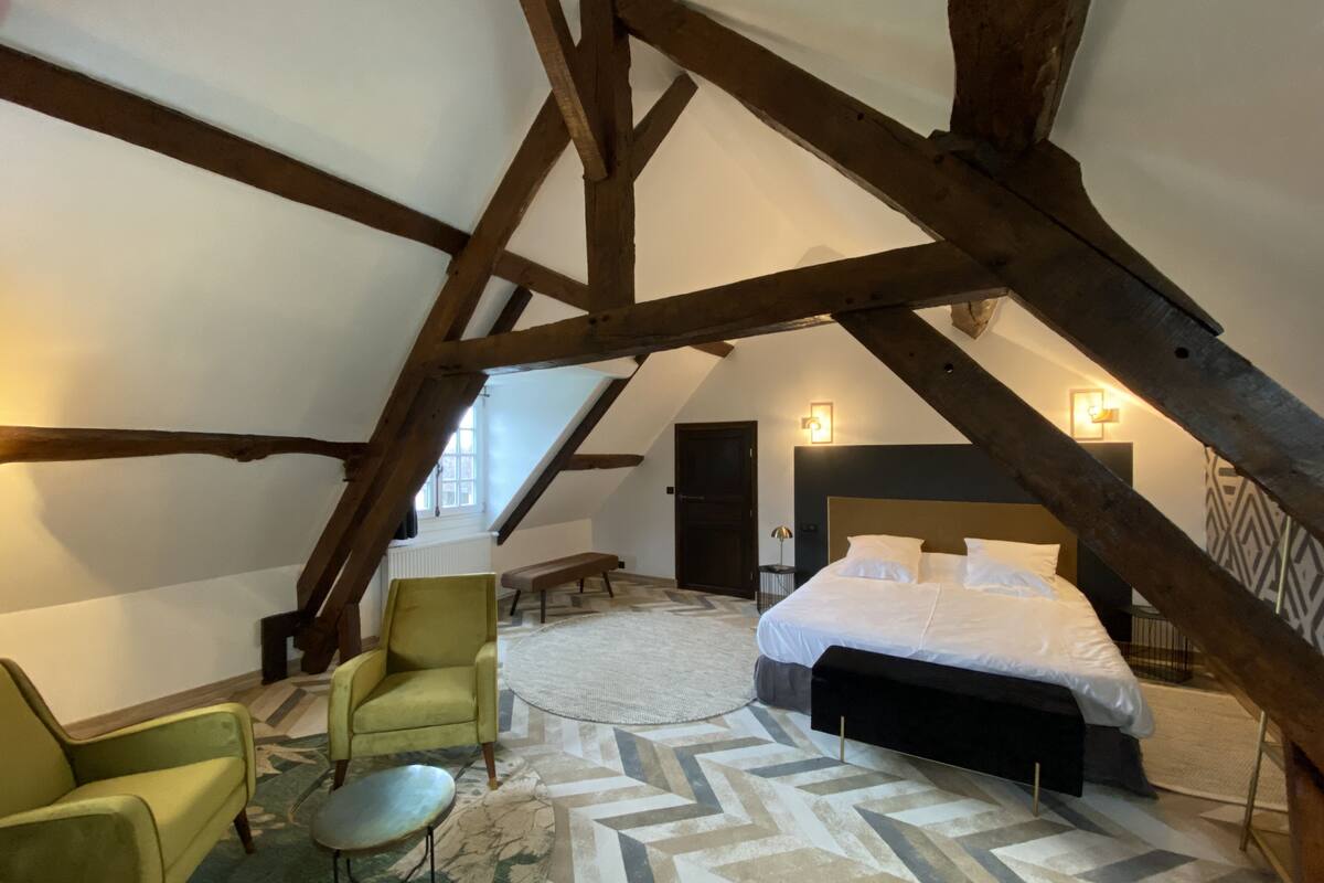 A bedroom with a large bed, a green armchair, a small round table, and a patterned rug. The room has exposed wooden beams and a sloped ceiling.