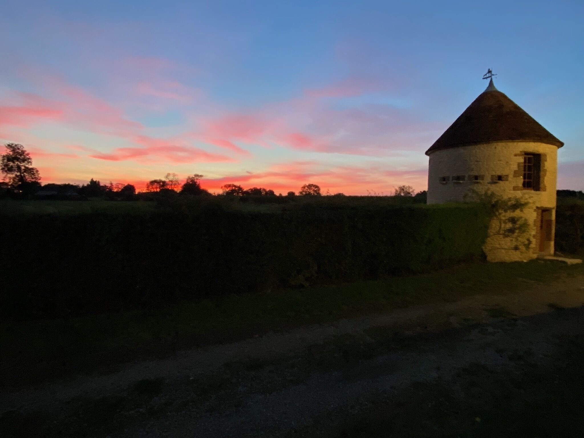 Un petit bâtiment isolé, doté d’un toit conique et d’une girouette, entouré de champs et bordé d’un chemin de terre.