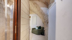 Un pasillo estrecho con una puerta de cristal, un muro con azulejos, un sofá verde con un cojín y una puerta blanca al fondo.