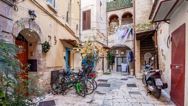 Una calle estrecha con pavimento de piedra, una puerta roja, una motocicleta y bicicletas estacionadas a un lado.