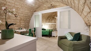Una habitación acogedora con techo inclinado, cama con ropa de cama verde, sofá verde, una pequeña mesa con sillas y una orquídea en maceta.