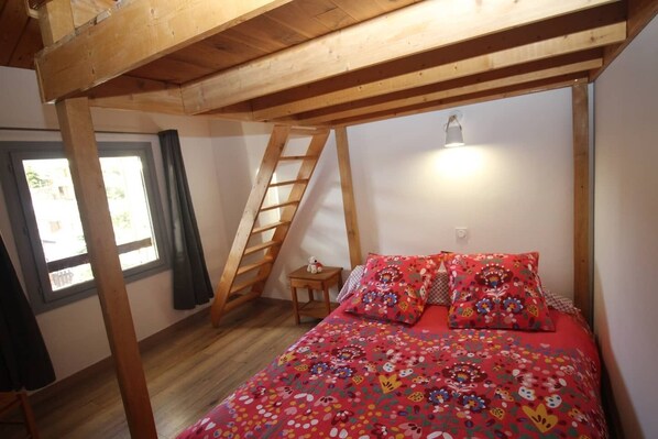 A bedroom with a wooden ceiling, a bed with floral bedding, a small table, a ladder, and a window with curtains.