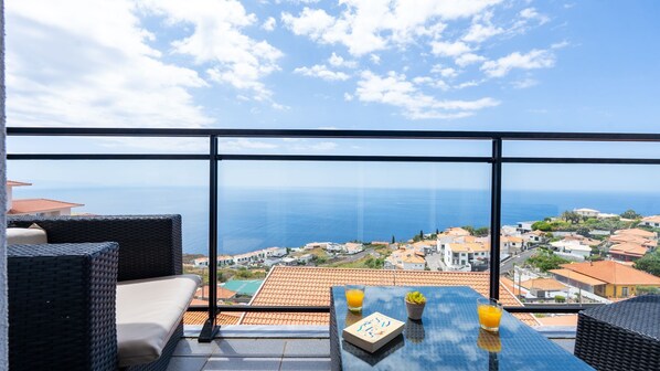 Un balcon avec vue sur la mer et sur la ville. On y trouve une table avec un livre et des verres de jus d’orange.